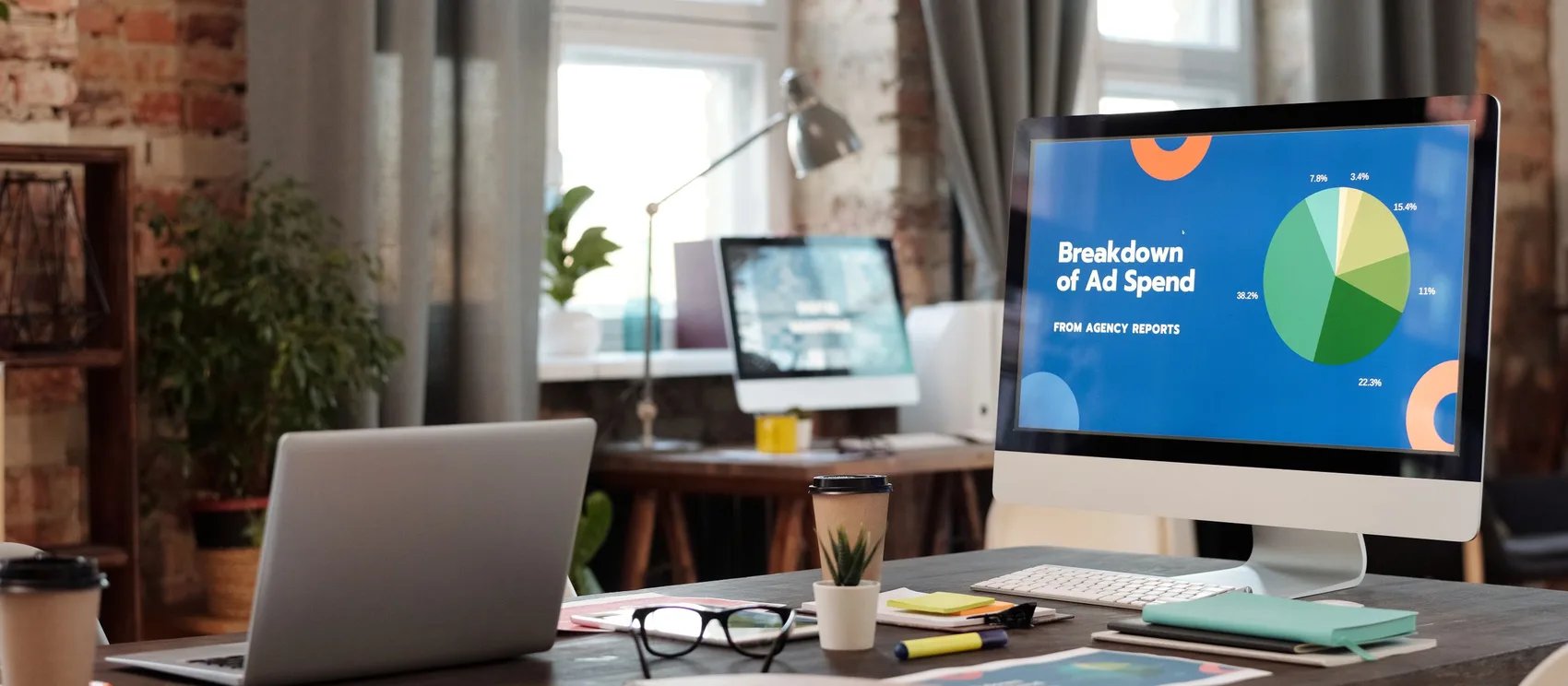 Monitor de escritorio mostrando un panel con la distribución de la inversión publicitaria en un gráfico de torta, en la oficina de una agencia con portátil, libreta y café.