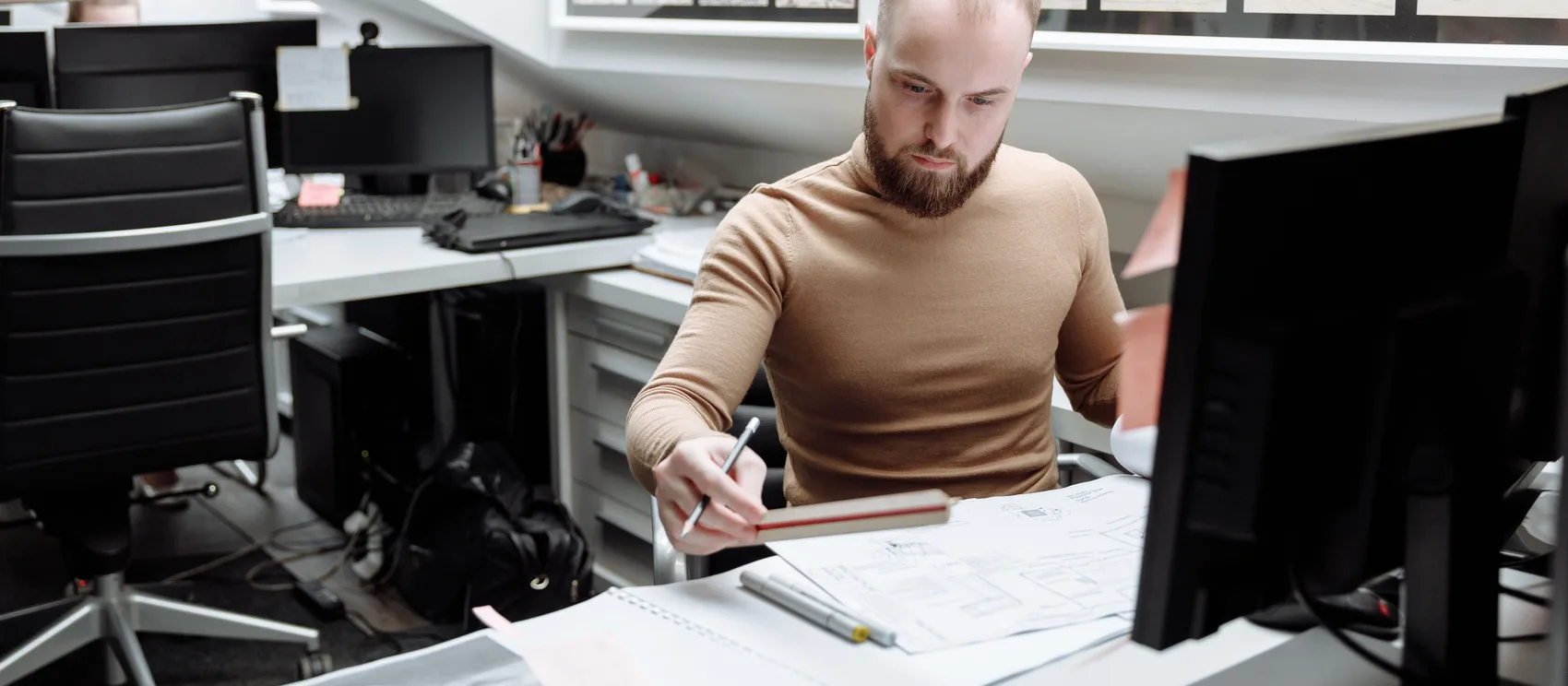 Arquitecto dibujando con regla y lápiz sobre planos arquitectónicos extendidos en una mesa de trabajo, con bocetos de arquitectura enmarcados en la pared de fondo